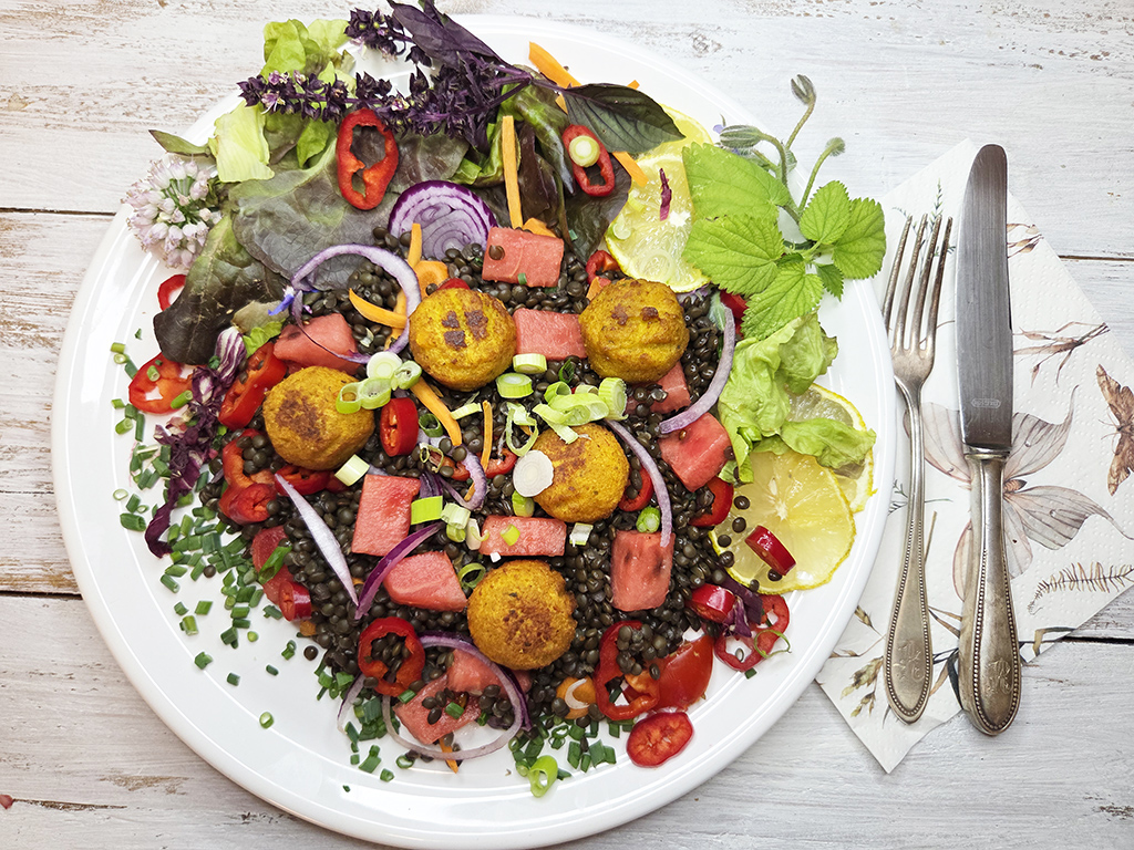 SOTO | Falafel Traditionell | Herzschatz Colorful plate of salad with falafel balls, watermelon cubes, red onion, peppers, greens, a lemon slice, and fresh herbs.