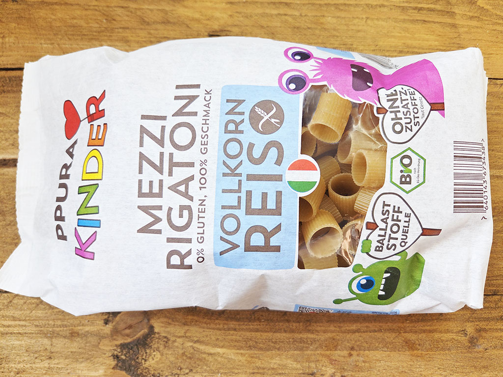 White bag of pasta on a wooden table, with colorful cartoon characters and a window showing pasta tubes on the packaging.