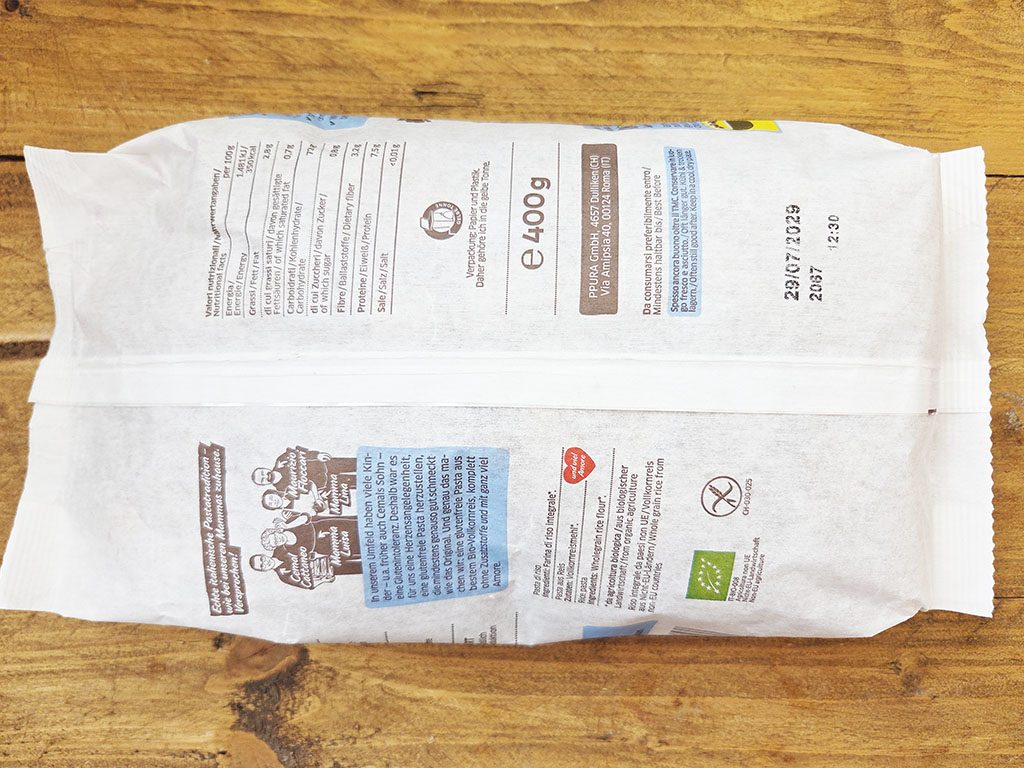 Two white paper food packaging sachets lying side by side on a wooden table, showing printed nutrition panels and logos.