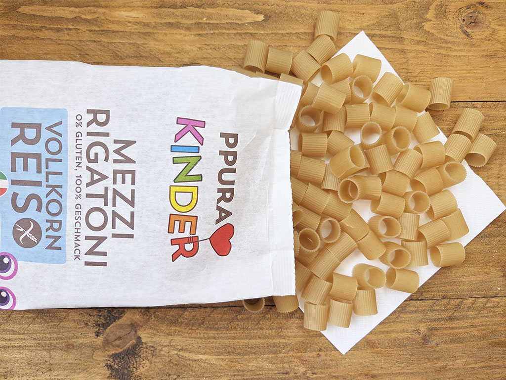 Uncooked small elbow macaroni spilling from a white paper bag onto a wooden table surface.