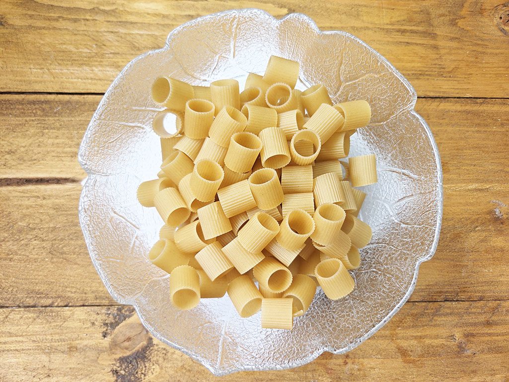 Uncooked rigatoni pasta in a clear glass bowl on a wooden table, ready for cooking.