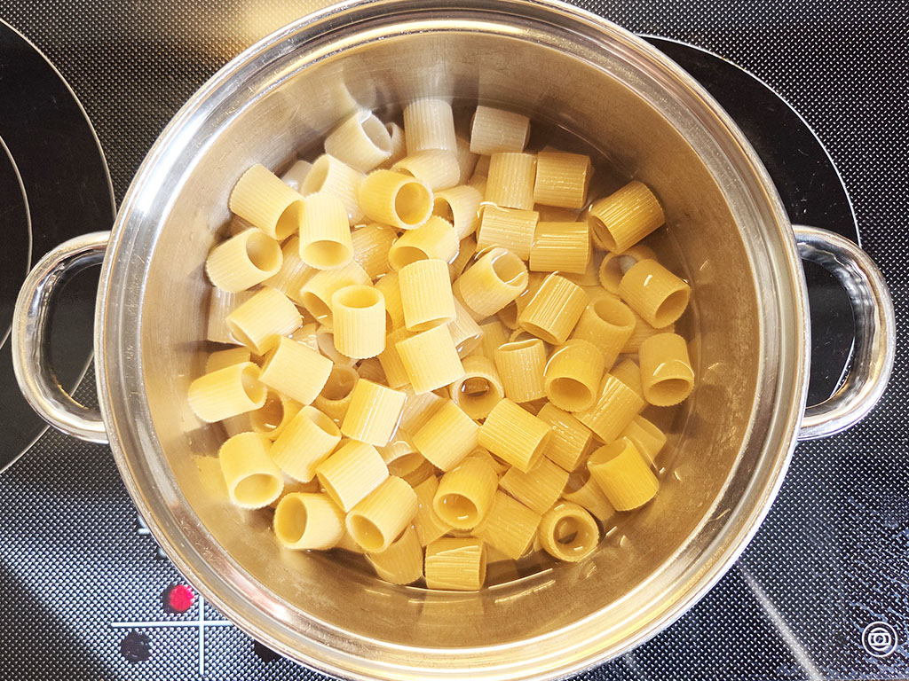 Uncooked rigatoni pasta in a stainless steel pot on a black electric stove, viewed from above.
