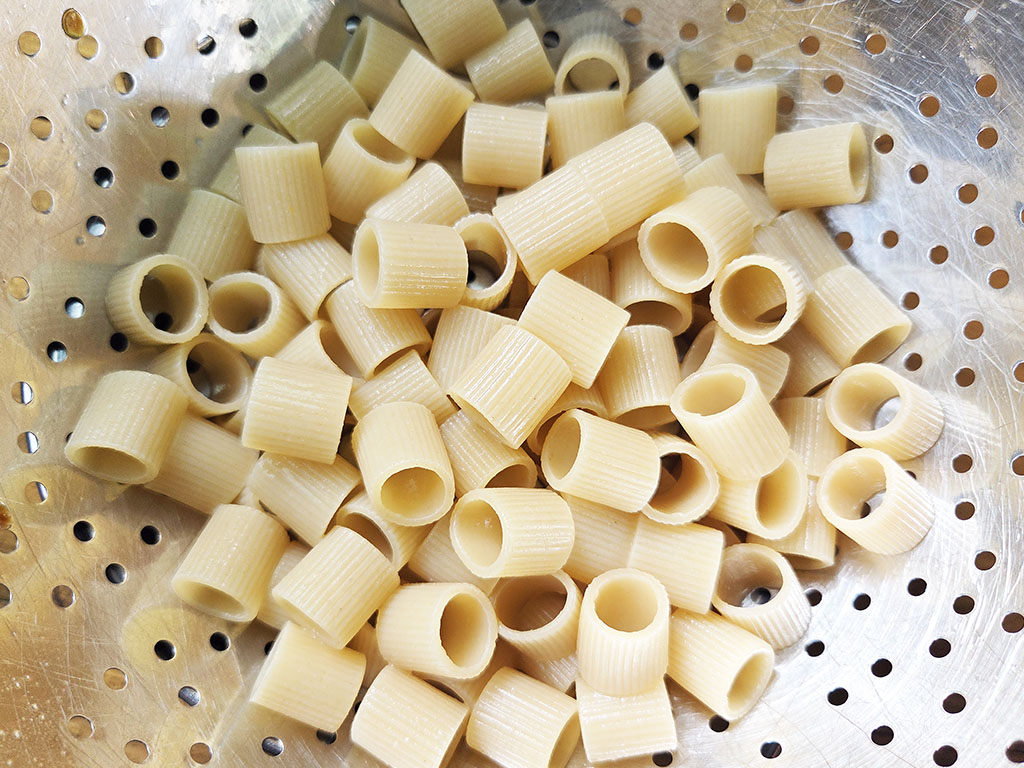 Uncooked ridged pasta tubes (small rigatoni) piled in a perforated metal colander/steam basket.