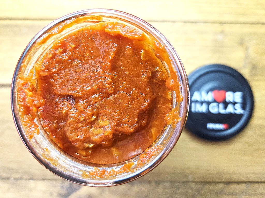 Open glass jar of thick orange-red sauce with the lid next to it on a wooden table.