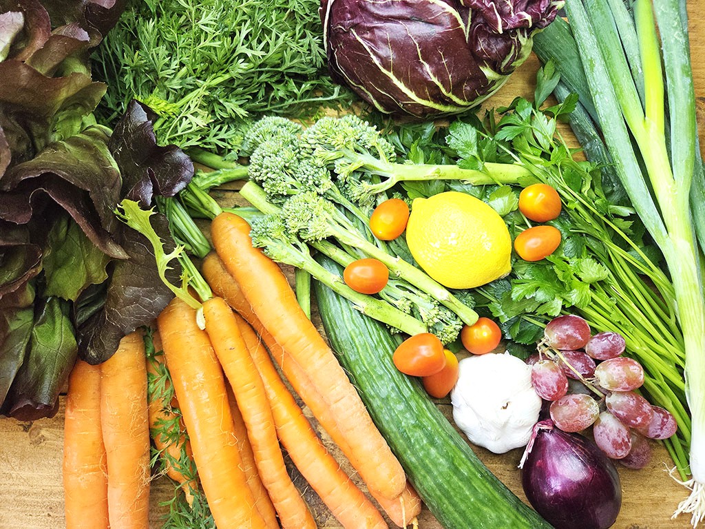 Colorful display of fresh produce on a wooden surface: carrots, cucumber, lemon, tomatoes, grapes, garlic, onion, leafy greens, and herbs.