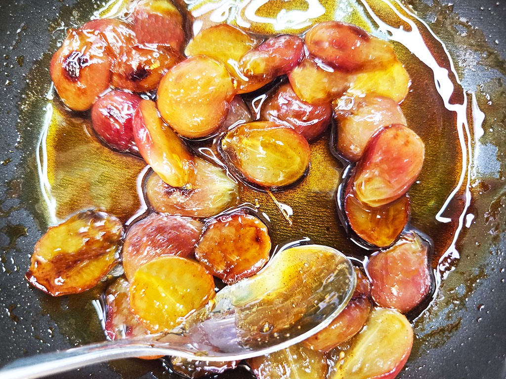 Sliced plums simmering in syrup in a frying pan with a spoon resting in the liquid, caramelized and glossy.