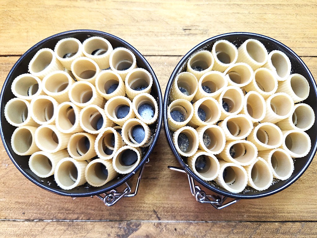 Two round pans filled with upright uncooked tubular pasta arranged in a circle on a wooden table surface.