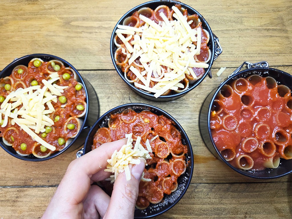 Top-down view of four bowls of pasta with red sauce and shredded cheese on a wooden table; a hand sprinkles cheese onto one bowl