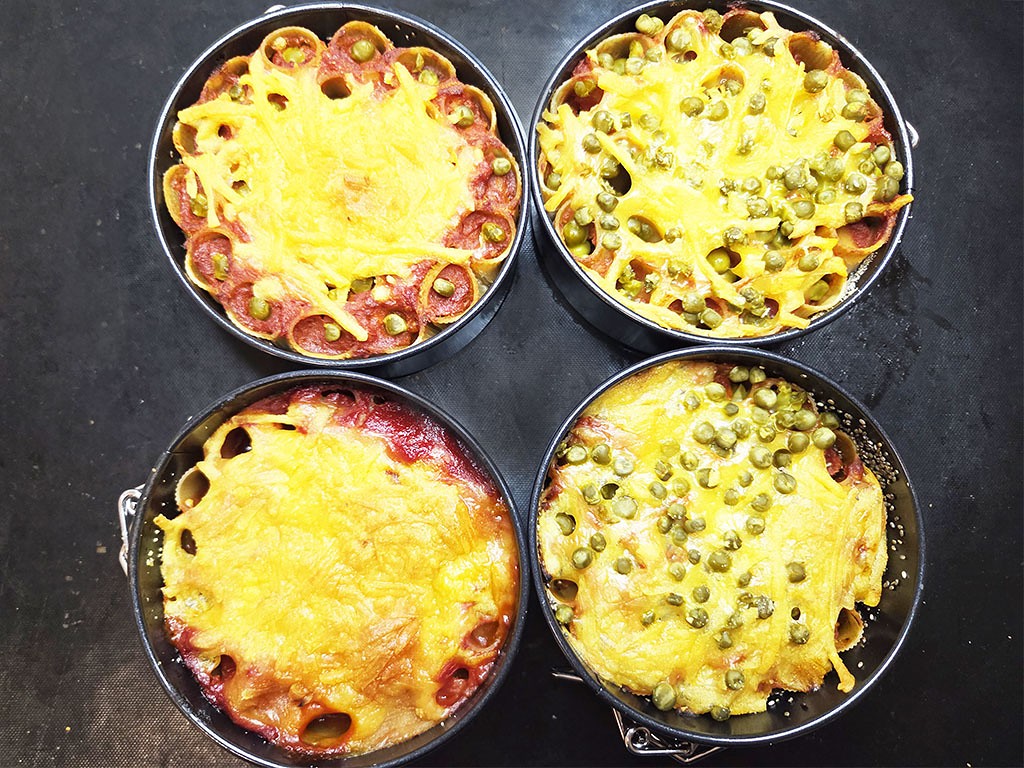 Top-down view of four mini pizzas in round pans, each with bubbling golden cheese and various toppings (pepperoni and peas).