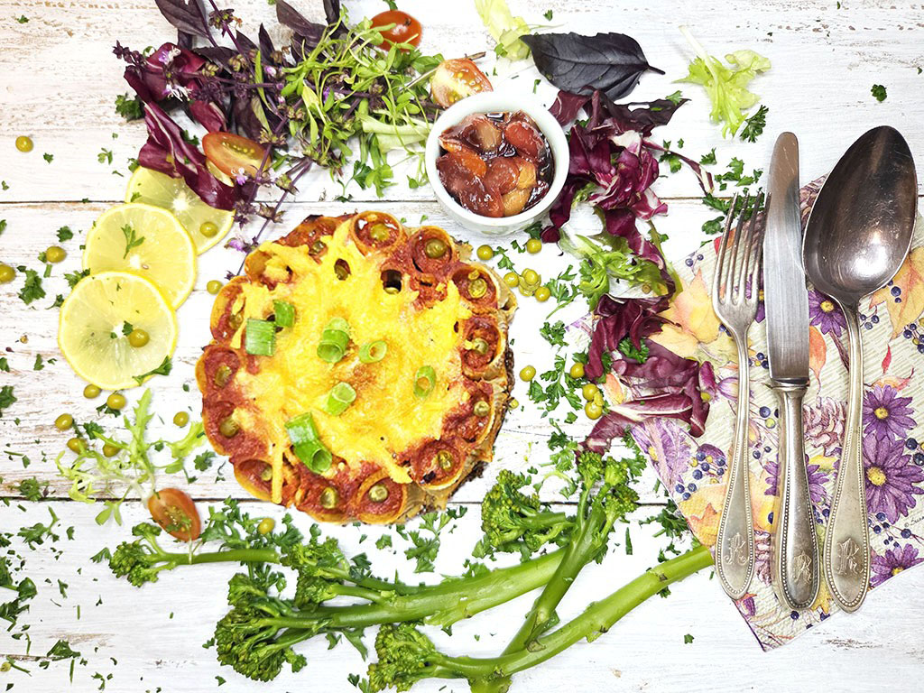 Cheesy baked dish topped with tomato slices and green onions, on a white wooden table with greens and lemon wedges nearby.