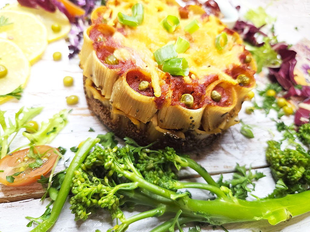 Slice of veggie casserole with olives, corn, and melted cheese on a crusty base, garnished with green onions, on a white rustic table with broccoli and lemon slices nearby.